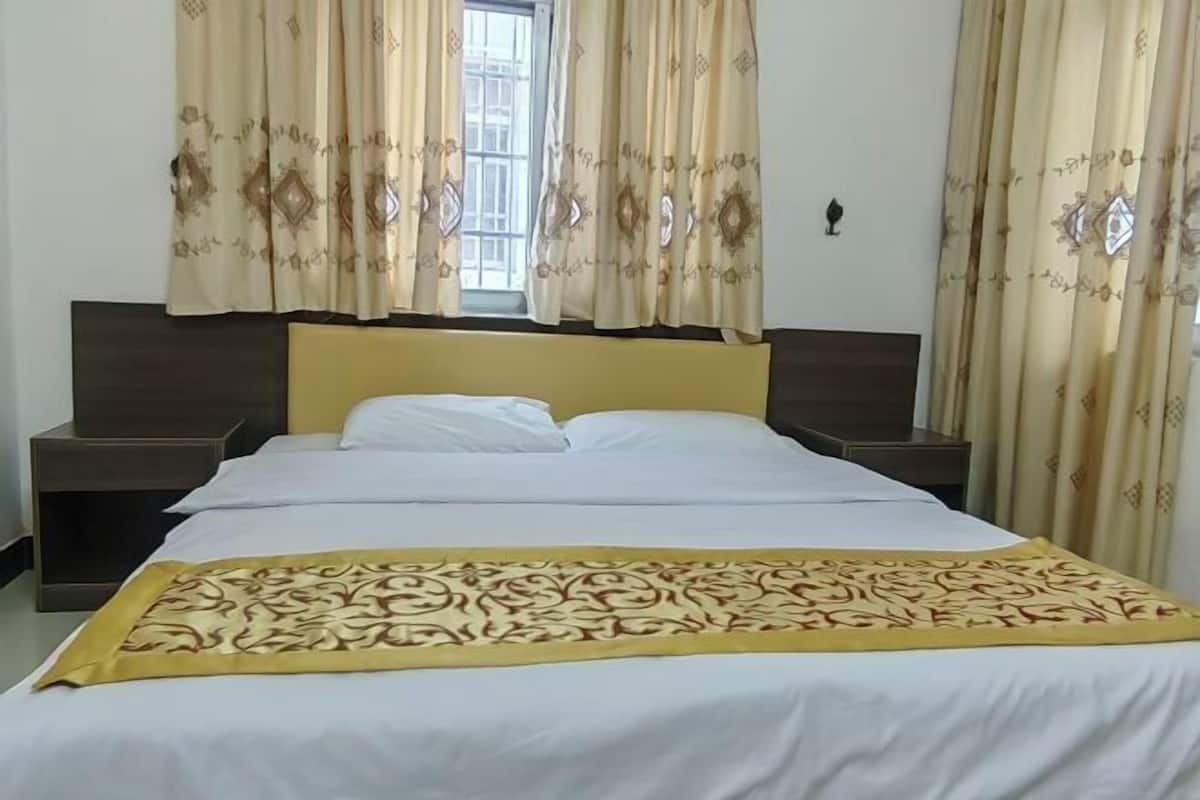 A hotel room with a bed, two bedside tables, a window with curtains, and a wall-mounted light.