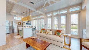 A spacious living area with a large window, a kitchen with a stainless steel refrigerator, and a wooden coffee table. - Shared Resort Style Pools, Boardwalk to the Beach, Golf Cart Access (Port Aransas)