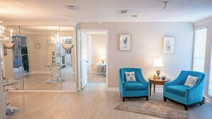 Een ruime woonkamer met een grote spiegel, twee blauwe fauteuils, een kleintje tafeltje met een lamp en een vloerkleed.