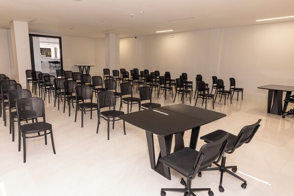 A conference room with multiple black chairs and tables, a white tiled floor, and a kitchen area in the background. - Riva Apart Hotel (Campo Mourao)