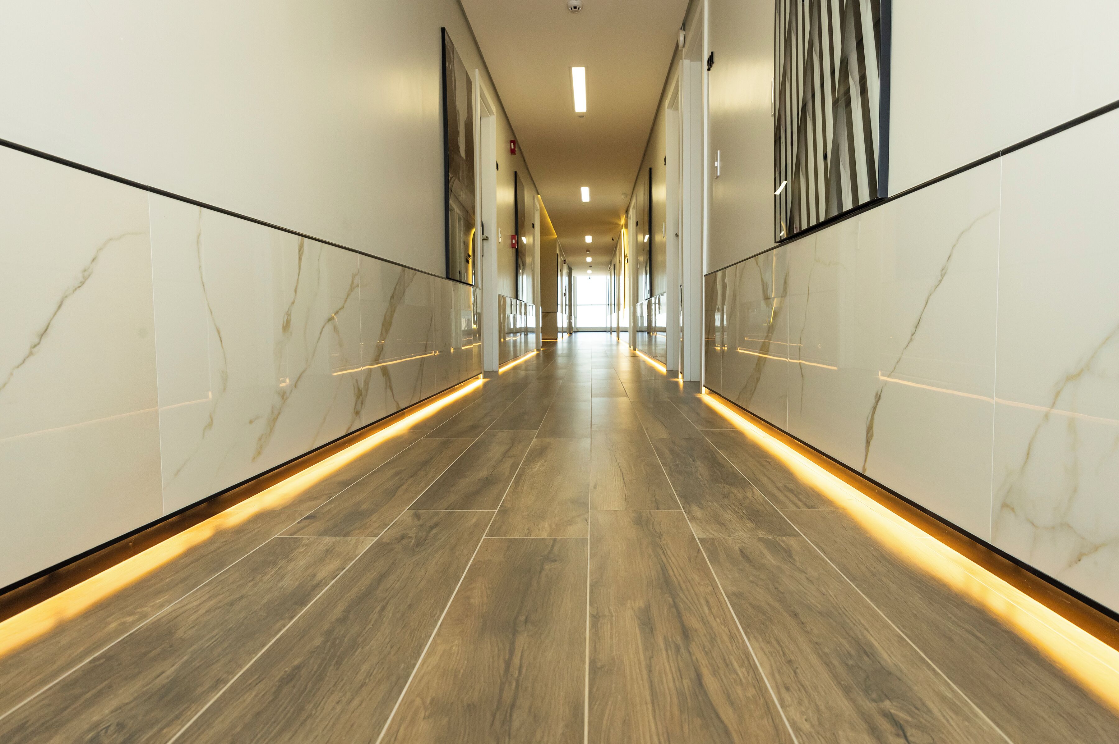 Un couloir moderne aux murs de marbre et au plancher en bois, qui mène à un espace lumineux et bien éclairé.
