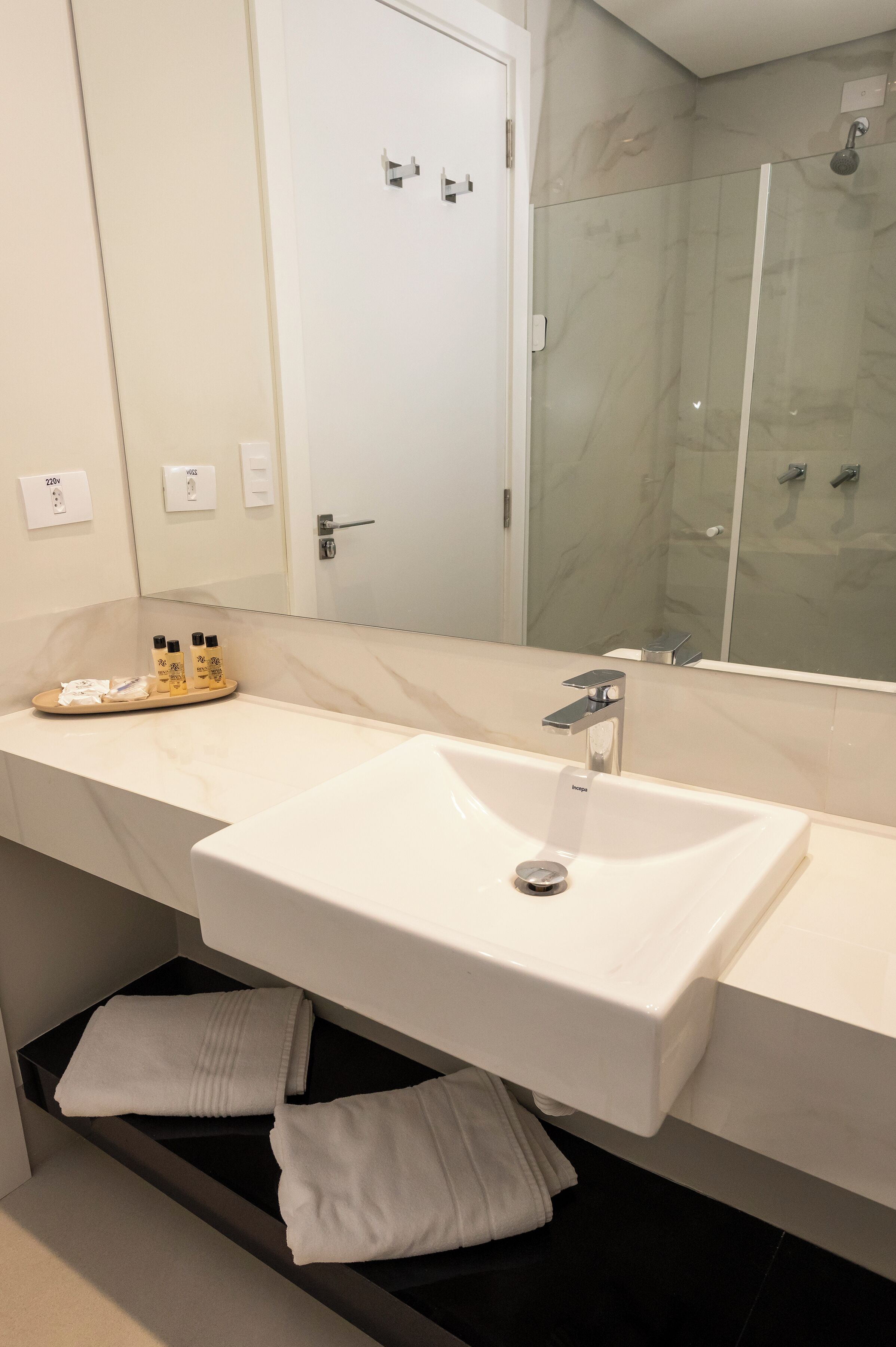 Un baño moderno con un lavamanos blanco grande, un espejo, cabina de ducha con mampara de vidrio y toallas dobladas con cuidado.