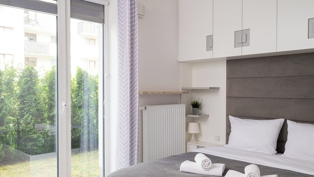 Ein modernes Schlafzimmer mit großem Fenster, einem Bett mit zwei gerollten Handtüchern, einem Nachttisch mit Lampe und Blick ins Grüne.