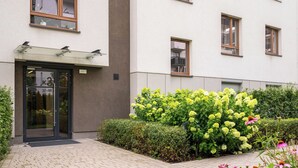 Ein Gebäudeeingang mit Glastür, gepflastertem Weg und einem Garten mit gelben Blumen.