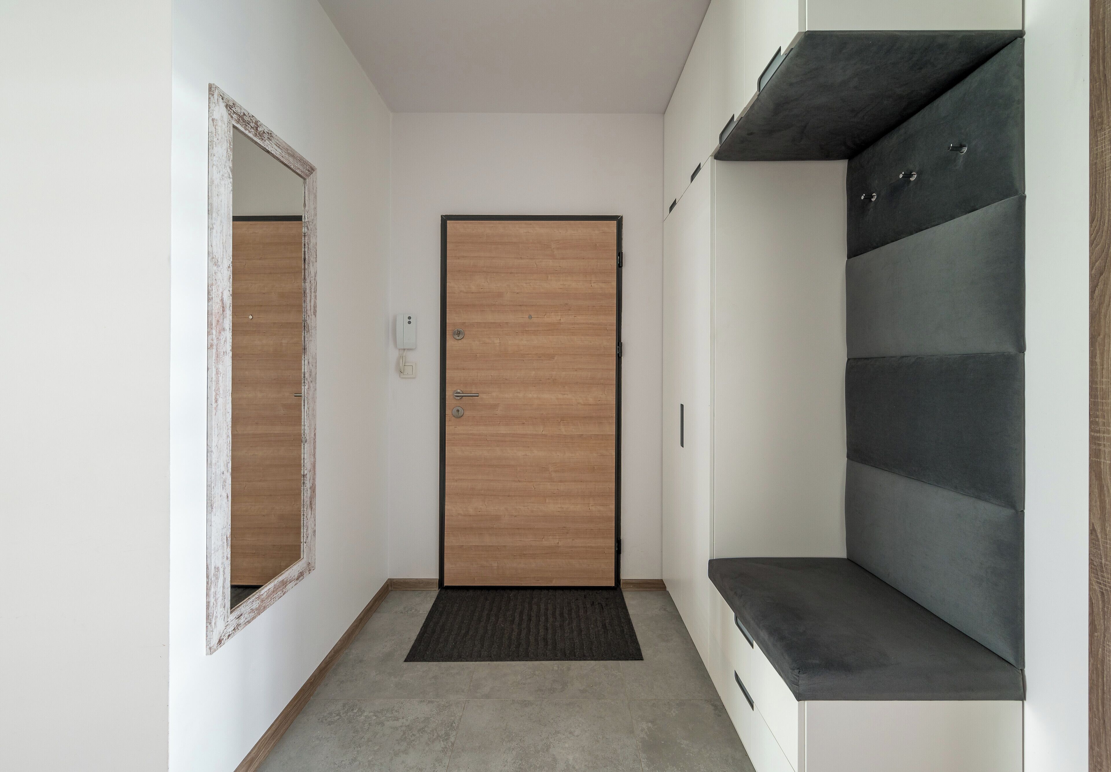 Un pasillo con una puerta de madera, un espejo y un banco gris.