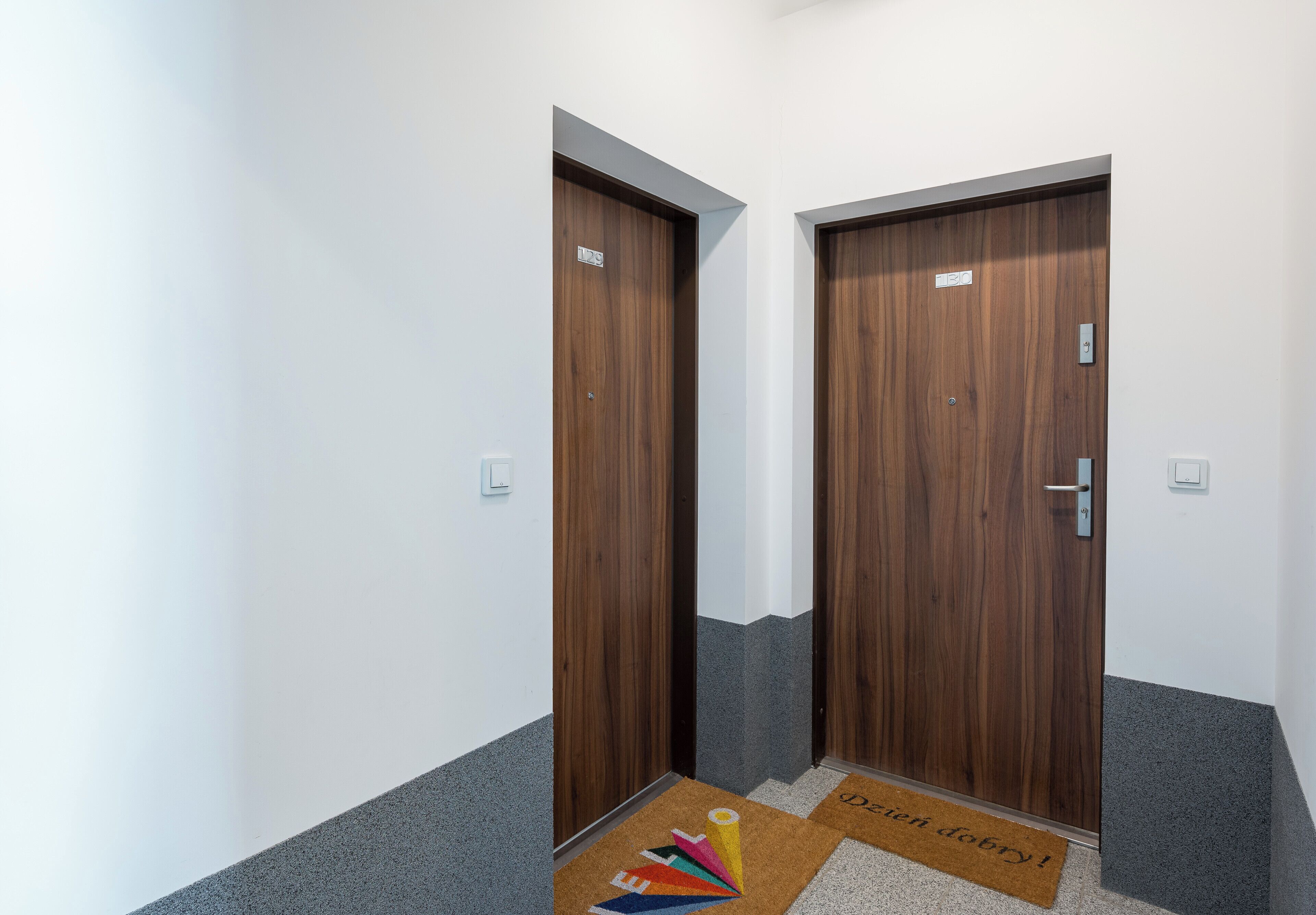Un couloir avec deux portes en bois, chacune surmontée d’une petite pancarte. On y trouve deux paillassons, l’un orné d’un motif géométrique coloré et l’autre portant l’inscription « Delici ! Delici ! ».