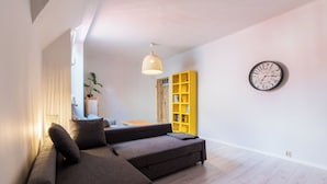 A modern living room with a sectional sofa, a yellow bookshelf, a round wall clock, and a hanging light fixture. - Głogowska Spacious Apartment (Poznań)