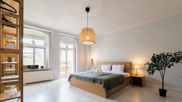 A bedroom with a wooden bed, a bedside table with a lamp, a potted plant, and a view through a large window. - Głogowska Spacious Apartment (Poznań)
