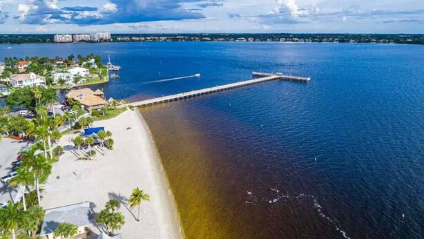 Ein Luftbild einer Küstenregion mit einer langen Pier, die sich ins Wasser erstreckt, einem Sandstrand und einem Wohngebiet mit Gebäuden und Palmen.