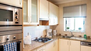 A compact kitchen with white cabinets, a stainless steel oven, a gas stove, a coffee maker, and a window with blinds. - Villa Emilly (Albufeira)