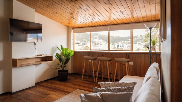 A modern living room with a wooden ceiling, a large window with a view, a TV mounted on the wall, and a seating area with a sofa and bar stools. - Suite de Lujo en Casa Patrimonial Reformada, en Zona Céntrica (Cuenca)