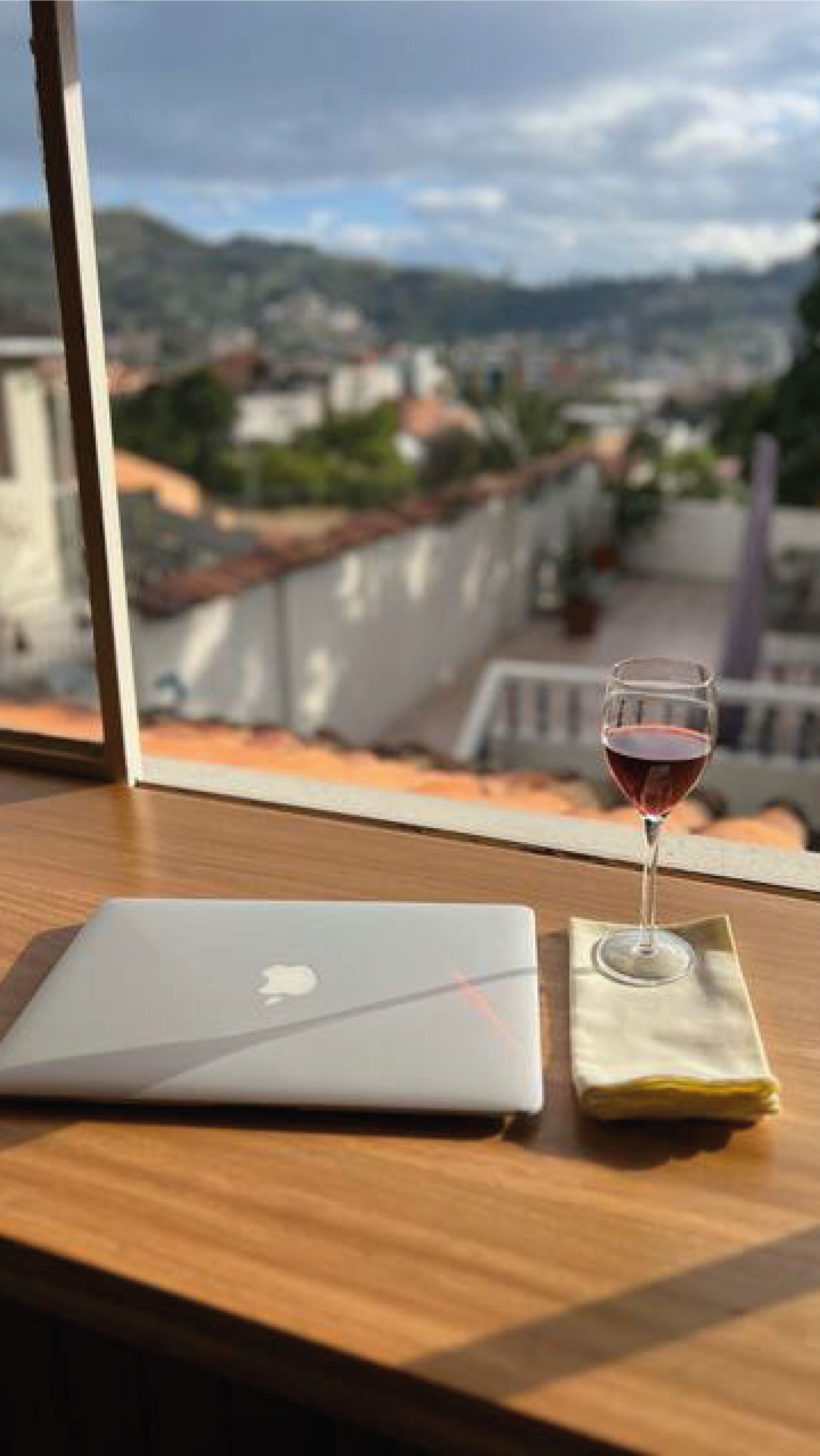 Un portatile su un tavolo di legno, con un bicchiere di vino rosso e una tovaglietta, con vista su una città che comprende edifici e colline.