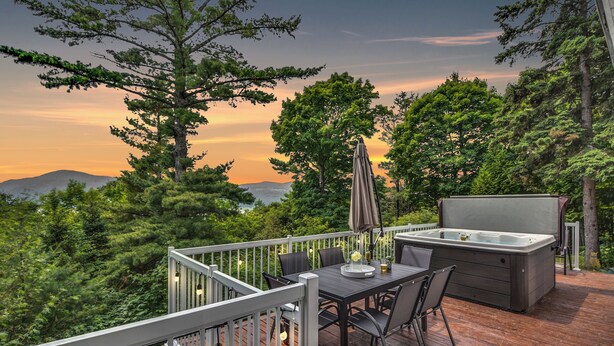Una terraza con mesa y sillas, un jacuzzi y vistas del bosque y las montañas circundantes.