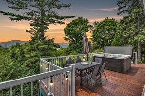 En terrasse med et bord og stole, et boblebad og udsigt over det omkringliggende skovlandskab og bjergene.