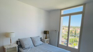 Ein Schlafzimmer mit einem großen Bett, zwei Nachttischen mit Lampen, einem Heizkörper und einem Fenster mit Blick auf eine Stadtlandschaft.