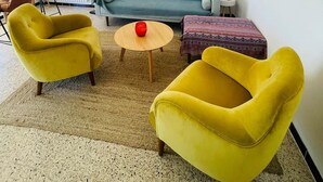 Ein Wohnzimmer mit einem blauen Sofa, zwei gelben Ohrensesseln, einem kleinen Holztisch und einem gemusterten Hocker.