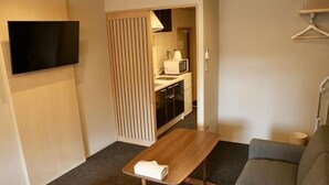 A hotel room with a bed, a wooden table with a towel, a grey sofa, a TV mounted on the wall, and a kitchenette in the background. - Hiz Hotel Gion-Shirakawa (Kyoto)