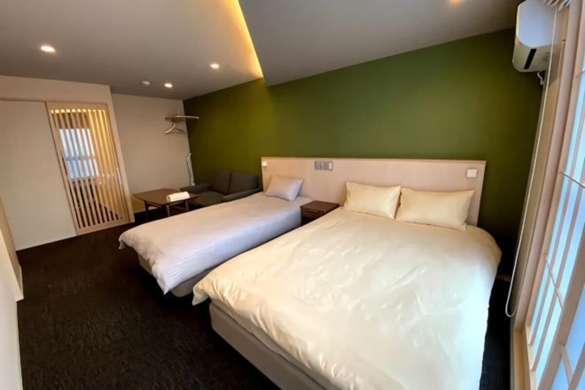 A hotel room with two beds, a sofa, a desk, and a window with blinds.