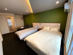 Habitación de hotel con dos camas, un escritorio, una silla, aire acondicionado colgante y una ventana con persianas.