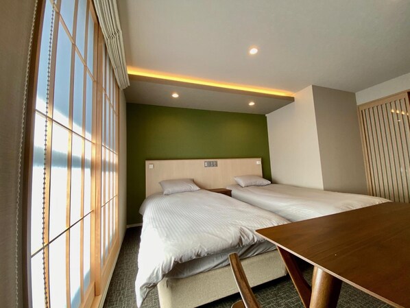 Une chambre d’hôtel avec deux lits, une table en bois et une vue par une fenêtre équipée de stores verticaux.