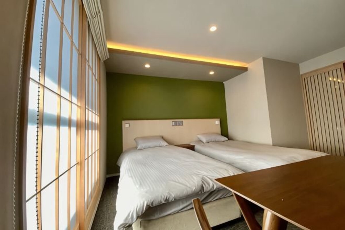 A hotel room with two beds, a wooden table, and a view through large windows.