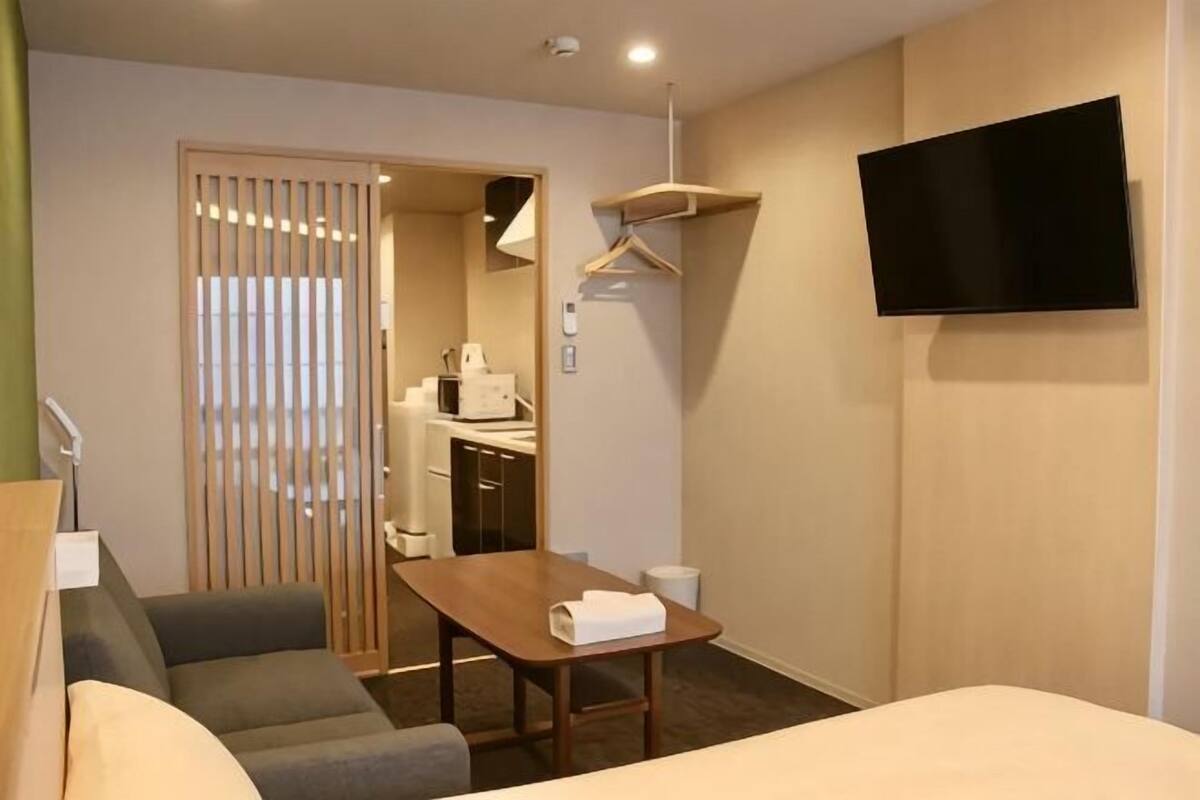 Ein Hotelzimmer mit einem Bett, einem Sofa, einem Tisch mit einem Handtuch, einem an der Wand befestigten Fernseher und einer durch eine Schiebetür einsehbaren Küchenzeile.