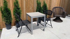 A patio with a table and two chairs, surrounded by a wooden fence and potted evergreen trees.