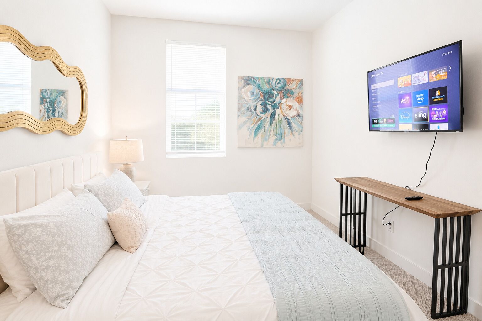 A bedroom with a bed, a TV mounted on the wall, a window with blinds, and a painting on the wall.