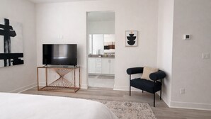 Una habitación de hotel moderna con una cama grande, un televisor sobre un mueble de madera, un sillón negro y una cocina visible a través de un vano abierto.