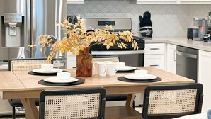 Una cocina moderna con electrodomésticos de acero inoxidable, una mesa de comedor dispuesta con platos y tazas, y un jarrón decorativo con hojas secas.