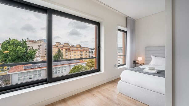 A modern hotel room with large windows, a bed, a bedside table, and a view of the cityscape.