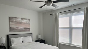 Quarto com cama, ventilador de teto, mesa de cabeceira com abajur, janela com persianas e um quadro na parede.
