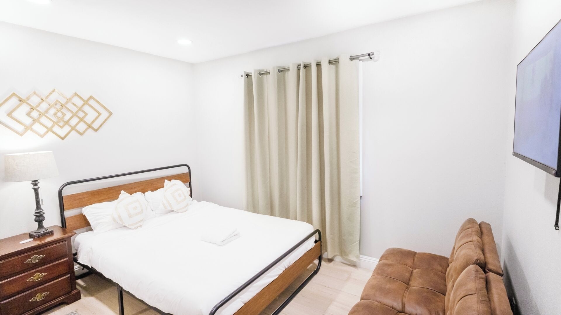 Une chambre à coucher avec un lit, une table de chevet, une lampe, un canapé marron et un téléviseur fixé au mur.