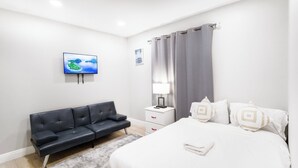 Una camera da letto moderna con un divano in pelle nera, un letto con biancheria da letto bianca e cuscini decorativi, una TV a schermo piatto montata a parete e un comodino con una lampada.