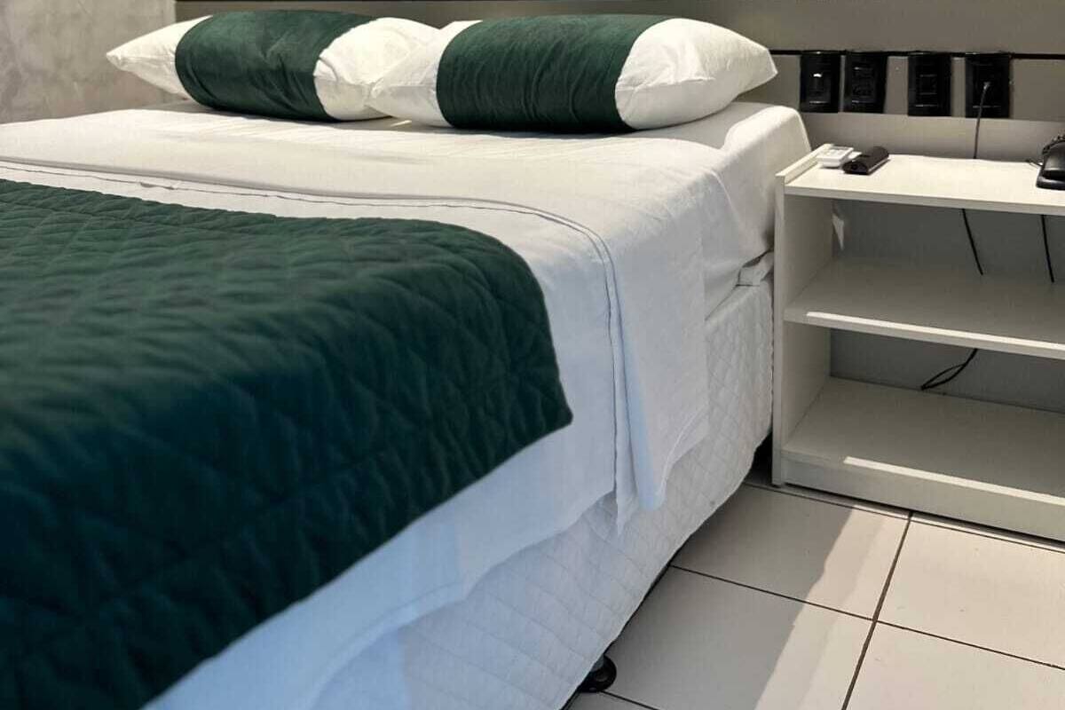 Una cama bien hecha con una colcha verde y blanca, un almohadón blanco y uno verde. Junto a la cama hay una mesita de noche blanca con un teléfono y una pequeña mesa.