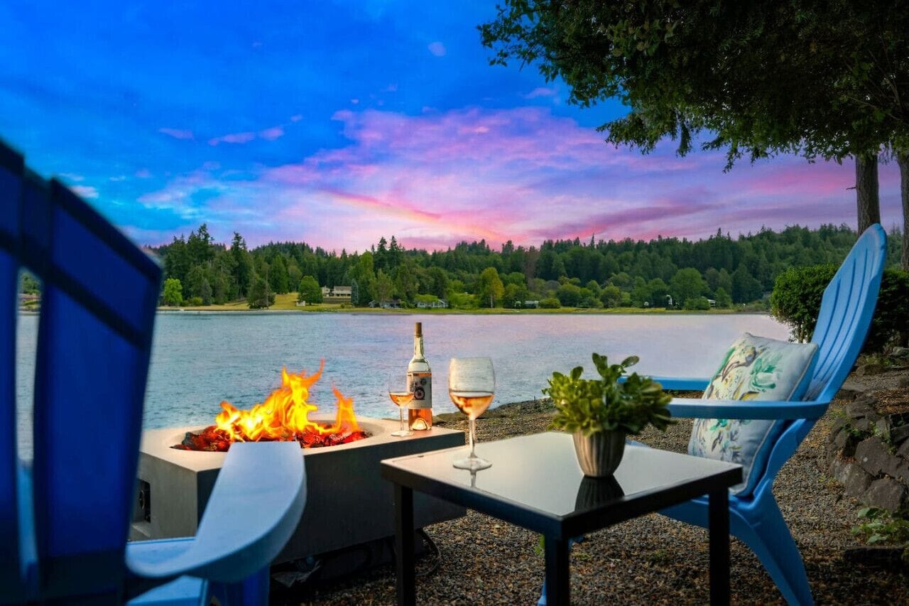 Zona de comedor junto al lago con un hogar, dos sillas, una mesa y una copa de vino.