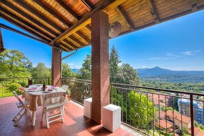 Ein Balkon mit Tisch und Stühlen, Blick auf Grünflächen und Berge, bewölkter Himmel.