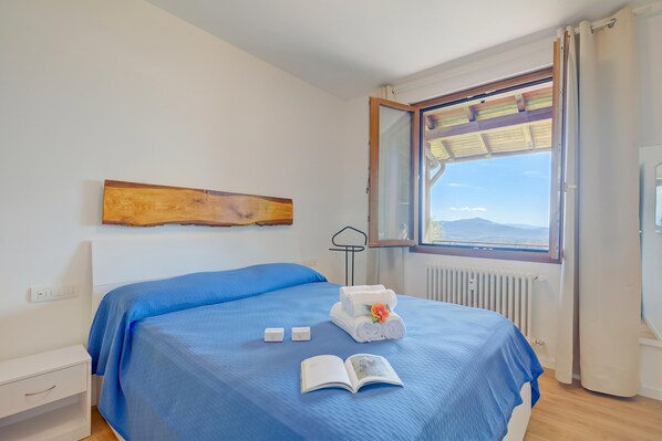 A neatly made bed with a blue bedspread, a rolled towel with a flower, and an open book on the bed.