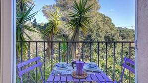 A balcony with a table set for two, surrounded by tropical plants and a view of a forested hill. - Studio Pinet - Near St Tropez - Pool, Ramatuelle, France (Ramatuelle)