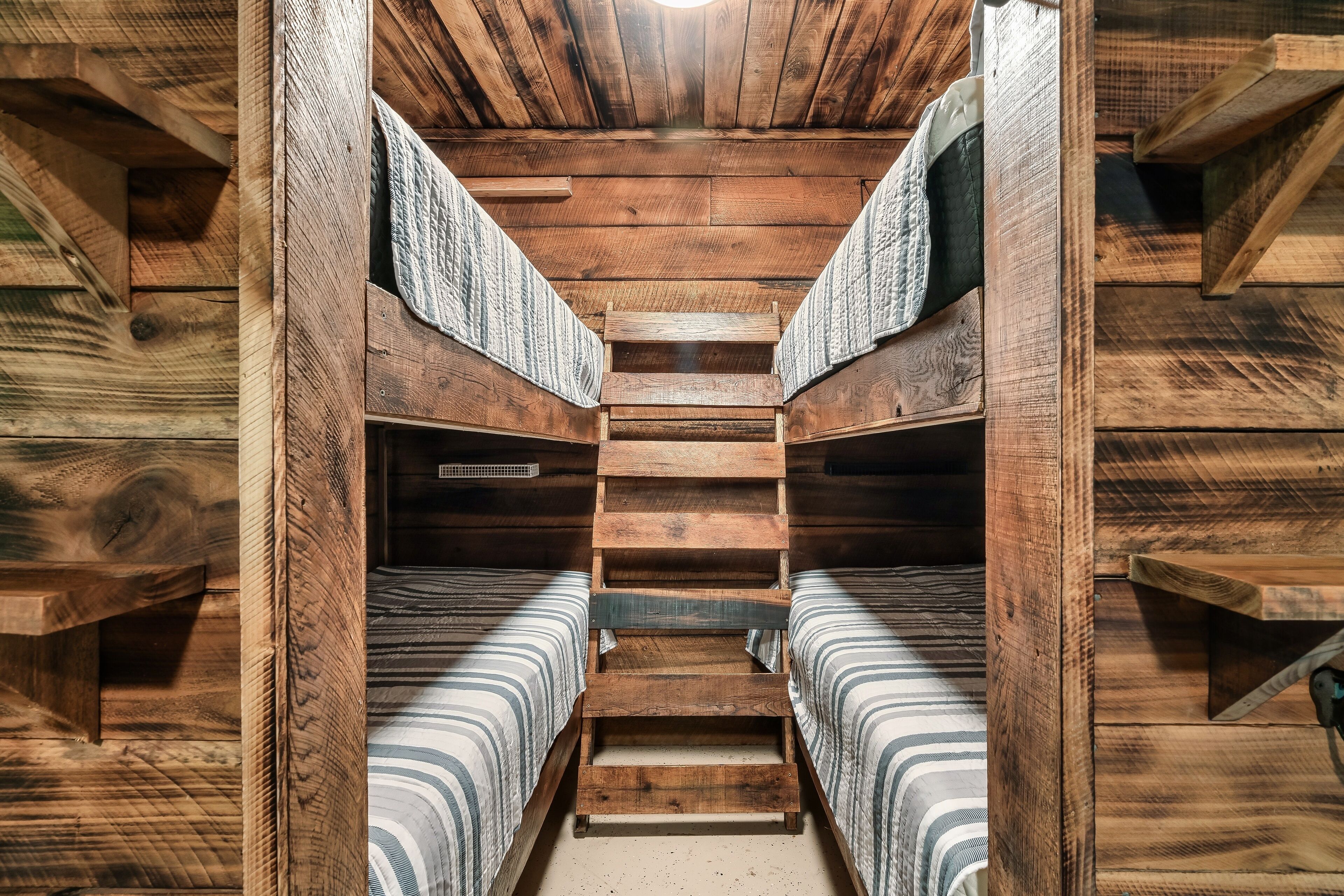 Interior de una cabaña de madera con una escalera central, dos literas y un estante pequeño.