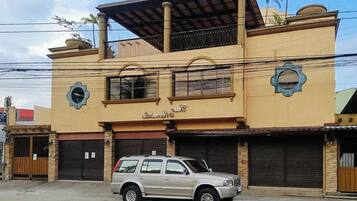 Un edificio de dos pisos con fachada color beige, un balcón en el nivel superior y un coche estacionado al frente.