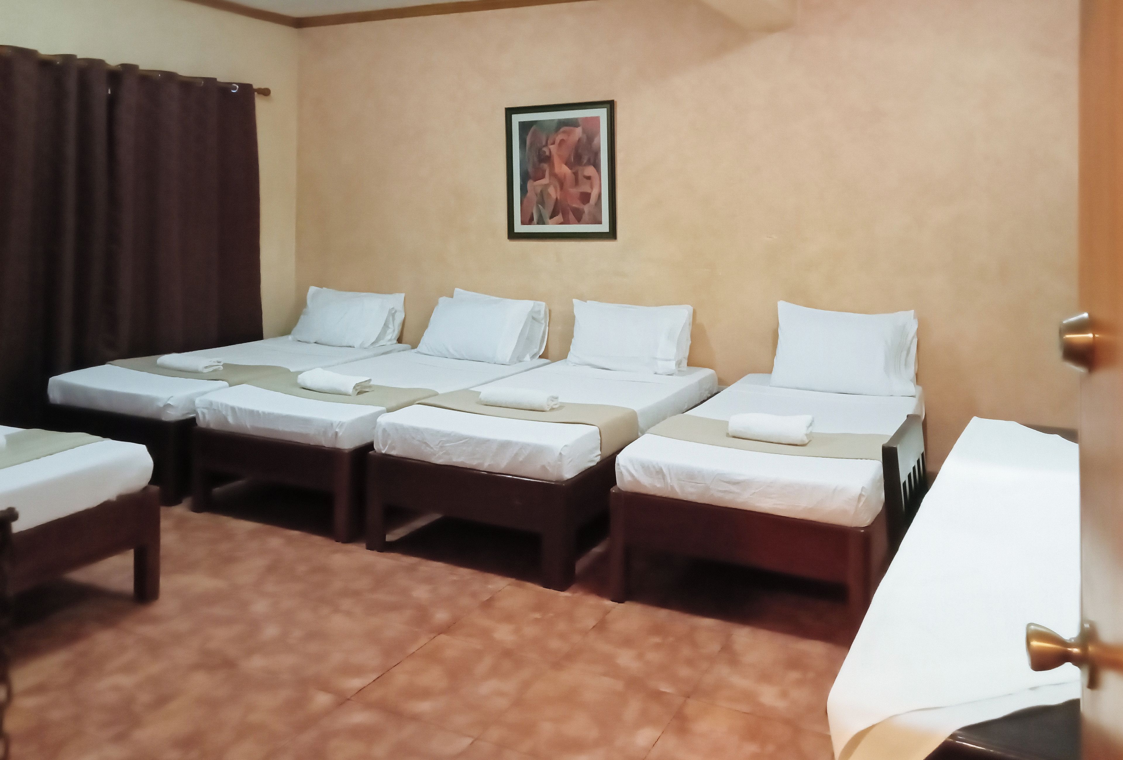 Habitación de hotel con cuatro camas individuales, una cortina marrón, un cuadro en la pared y una puerta con pomo de latón.
