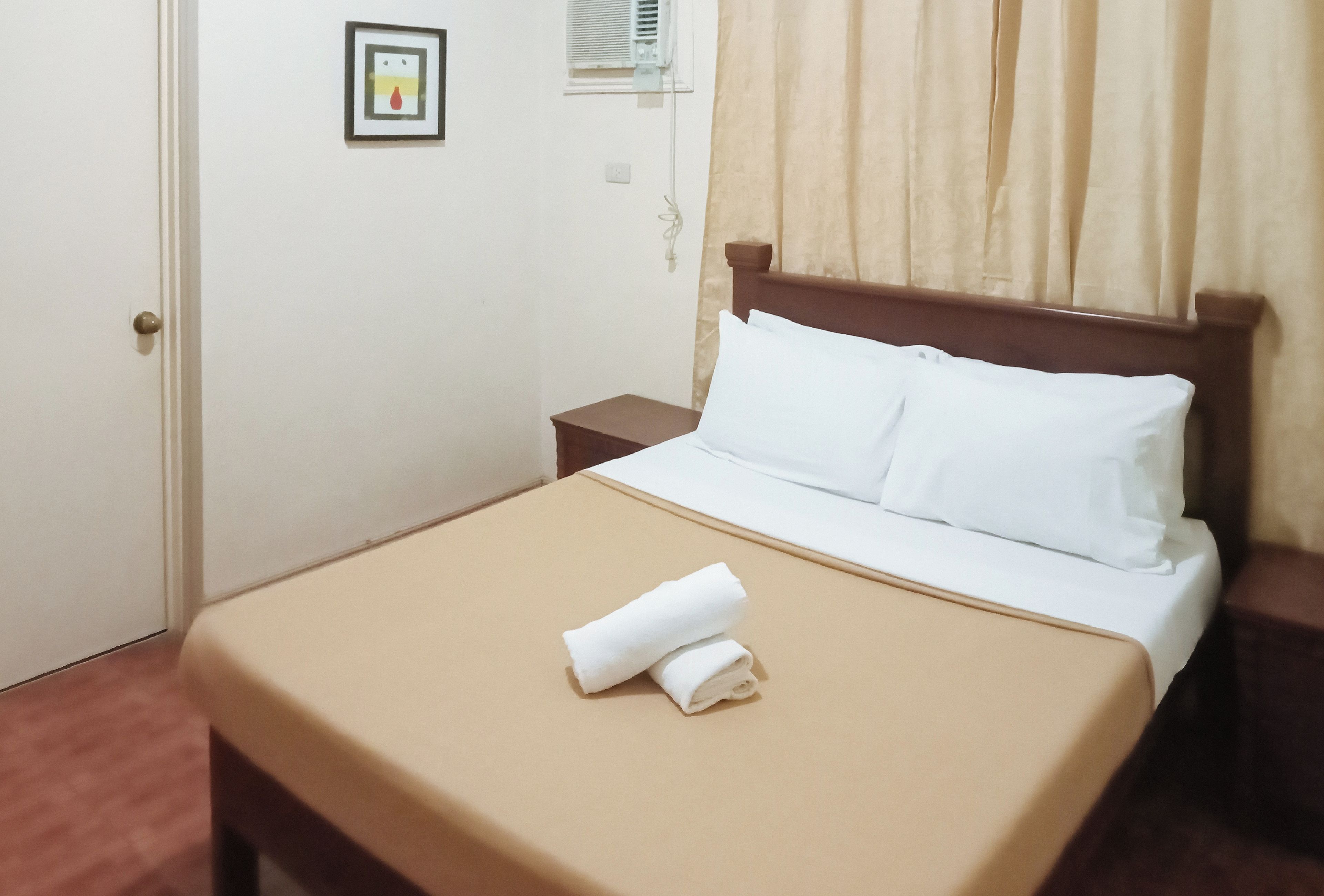 Una cama bien hecha con sábanas blancas y una colcha marrón, una mesita de noche pequeña y un equipo de aire acondicionado instalado en la pared.