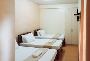 A hotel room with three single beds, each with a wooden headboard, white bedding, and a rolled towel. There is a wardrobe with two doors and a small shelf above it.