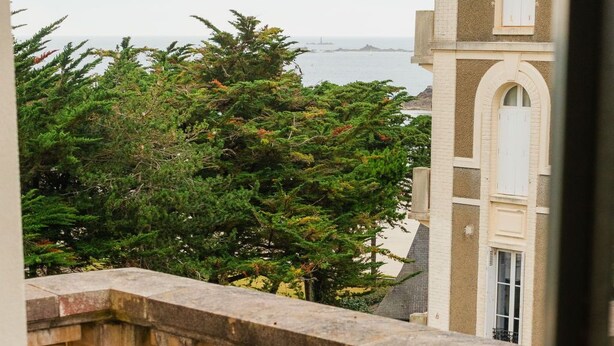 Un balcón con vistas a un edificio y árboles. Hay una silla verde y una mesa con un jarrón de plantas secas.
