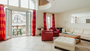 Un salón espacioso con un ventanal grande, cortinas rojas, un sofá, un sillón rojo, una mesa de centro de madera y una mesita auxiliar.