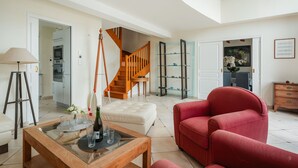 Amplia sala de estar con una mesa de centro de madera, sillones rojos, una mesa con cubierta de vidrio y bebidas, una escalera y una cocina al fondo.