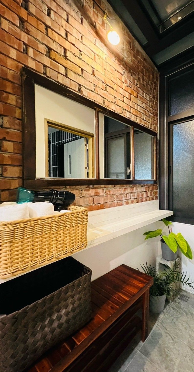 Une salle de bain avec un mur en briques, un comptoir blanc, un miroir, un lavabo, un panier, un banc et une plante en pot.