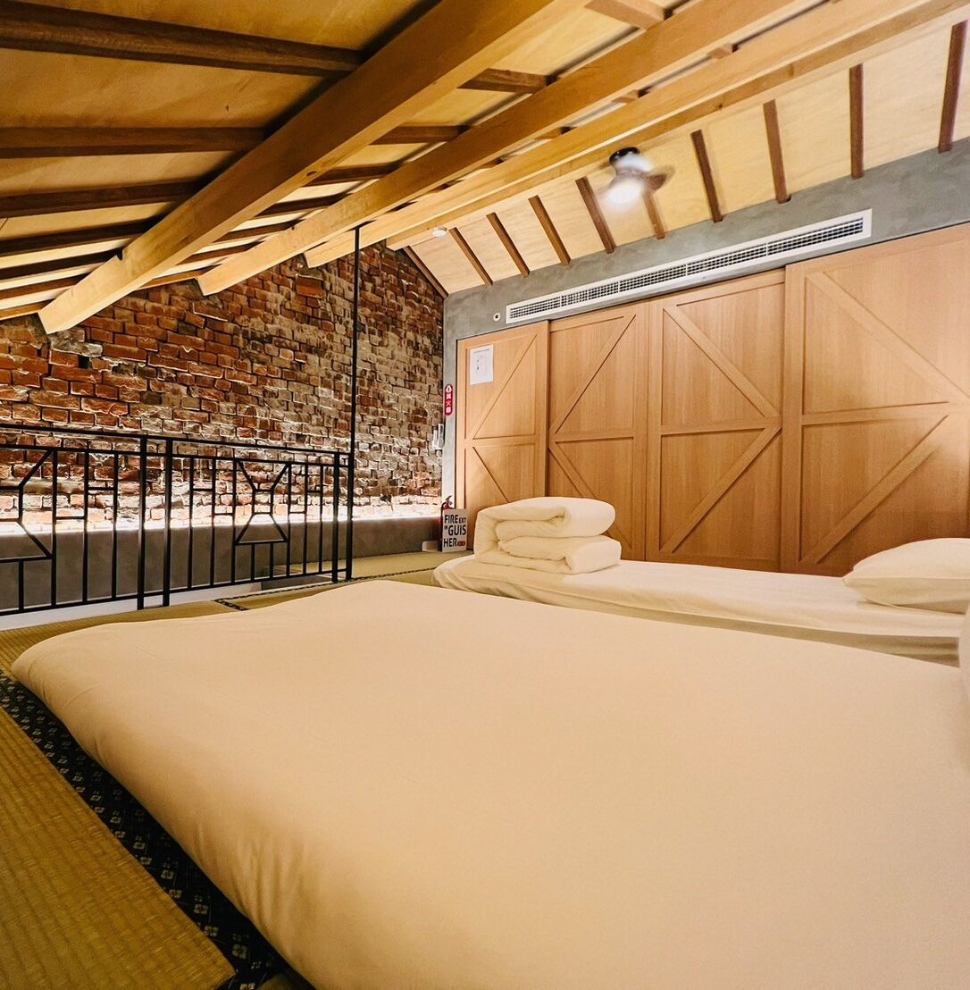 Habitación de hotel con pared de ladrillo, vigas de madera en el techo, cama con ropa blanca, una pila de toallas y una pequeña zona de estar con barandilla de metal.