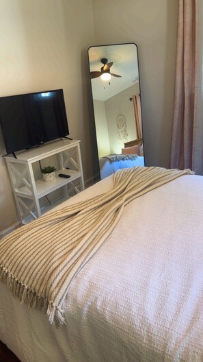 Un dormitorio con una cama, un televisor sobre un soporte blanco, un espejo y un ventilador de techo.